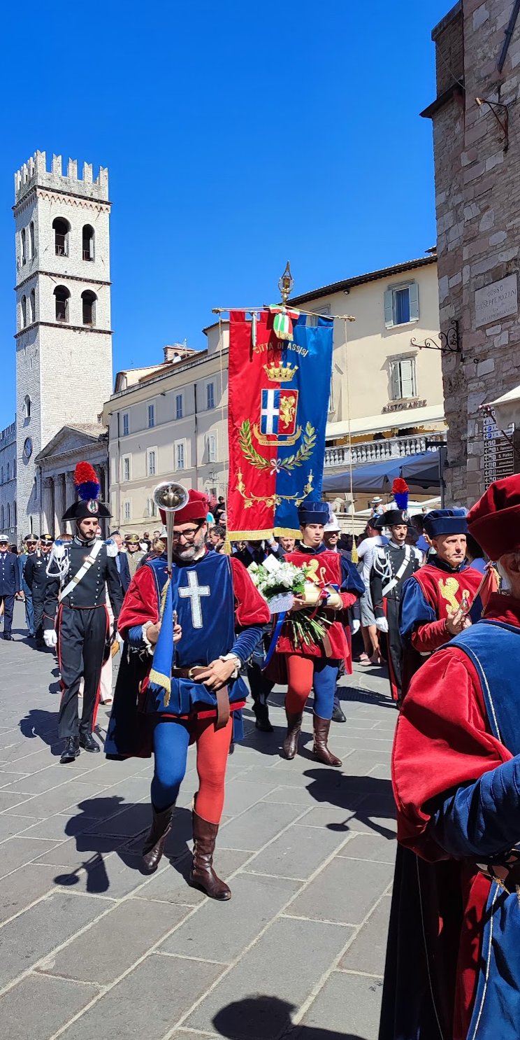 A civic procession in historic costume crossing a piazza in Assisi, with a stone tower in the background