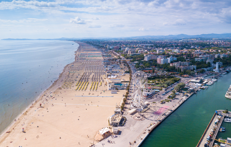 Aerial view of a crowded Adriatic beach in mid-August, with rows of umbrellas stretching along the shoreline next to a harbor