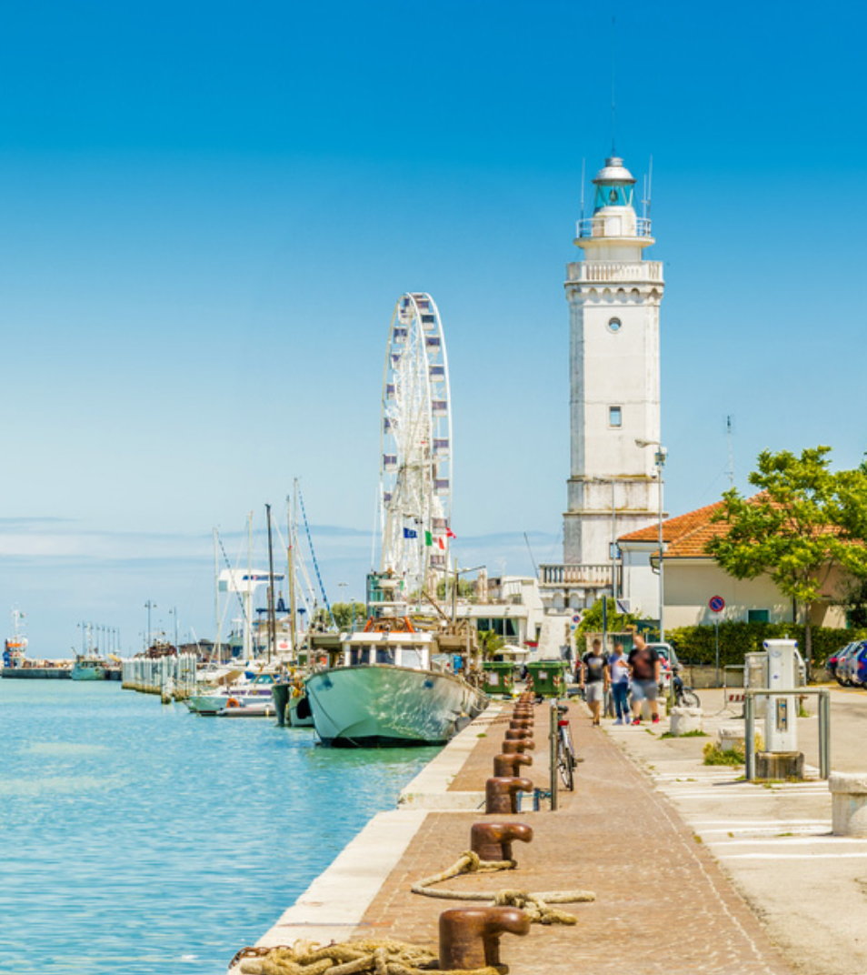 A historic white lighthouse and a Ferris wheel at the entrance to an Adriatic harbor, with fishing boats moored along the quay