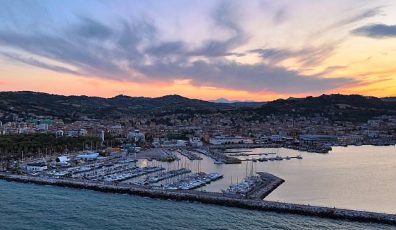 Aerial view of an Adriatic marina and harbor town at sunset, with hills rising behind the rooftops