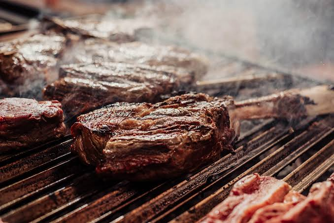 Close-up of meat grilling over an open flame at a sagra cooking station, smoke rising from the grate