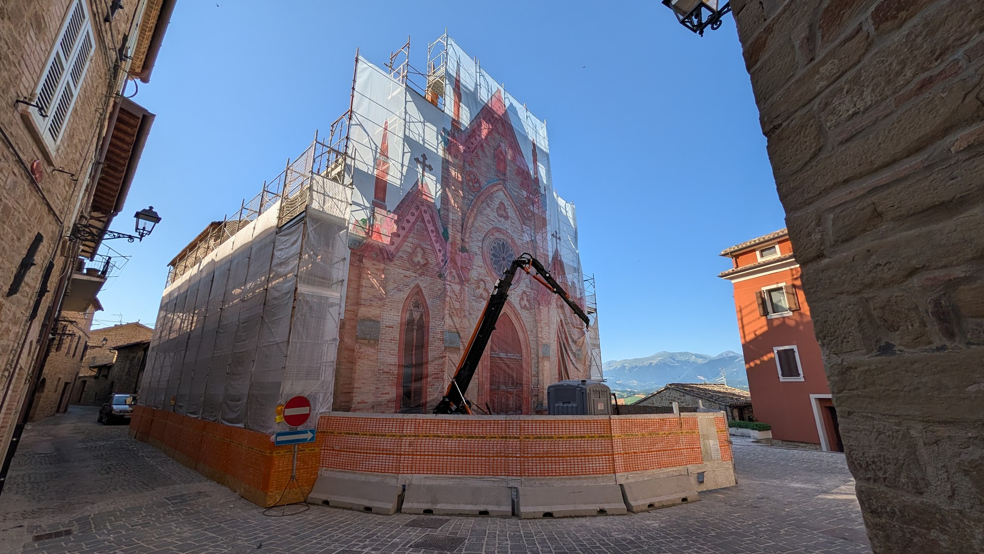 A Gothic church facade in the Marche under seismic reconstruction, its original stonework printed on the scaffolding wrap, mountains in the background