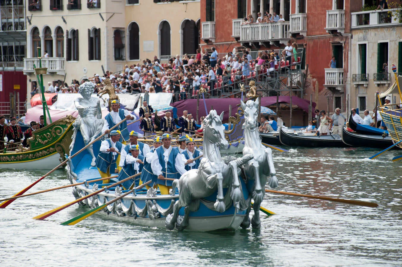 A ceremonial Regata Storica boat decorated with silver horse figureheads passing crowded grandstands on the Grand Canal